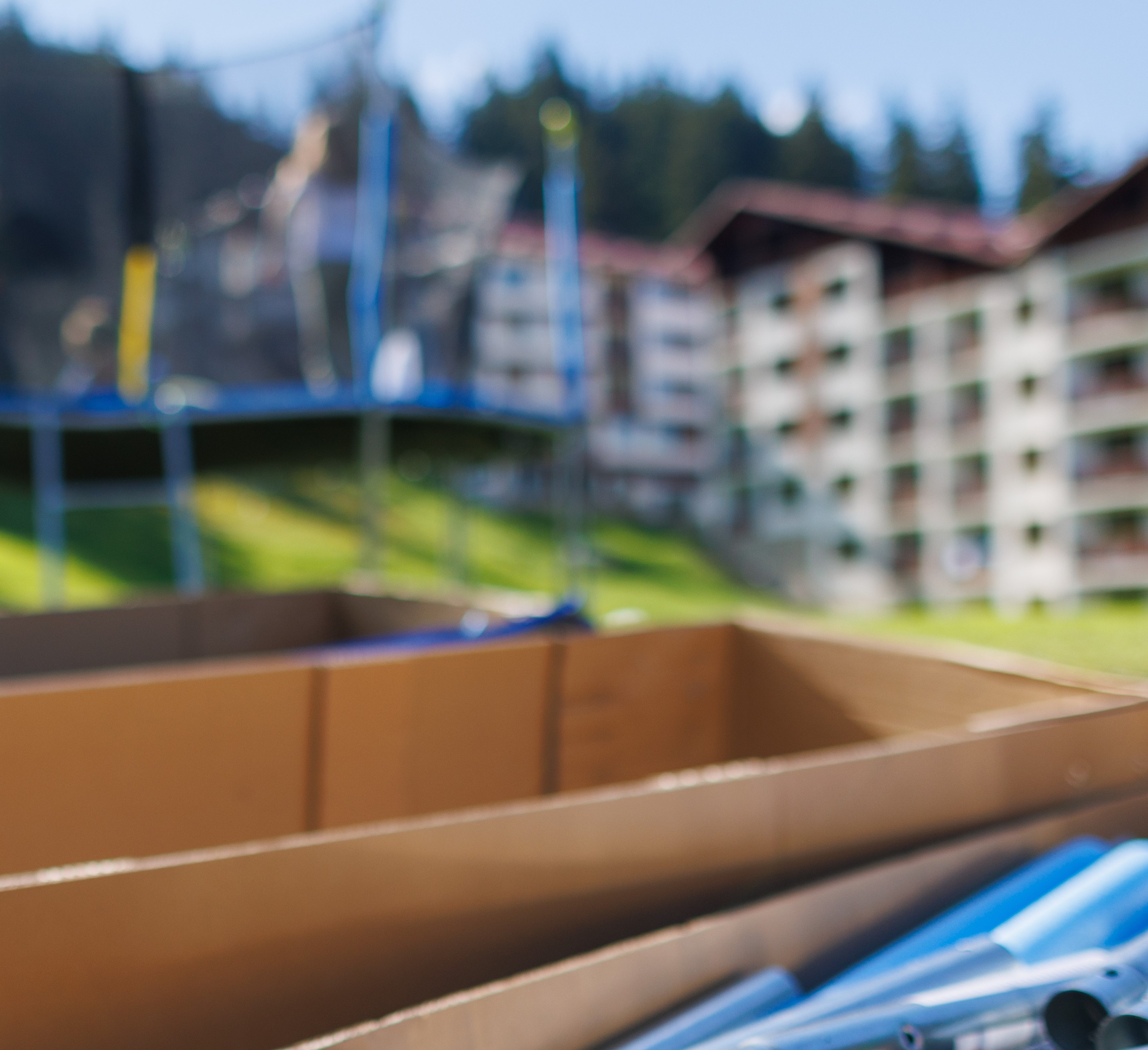 Boxes with iron beams to assemble a trampoline as in the background next to people assembling a trampoline Large cardboard boxes with iron beams and other kit to assemble a netted trampoline as in the background next to people assembling a trampoline on a backyard lawn